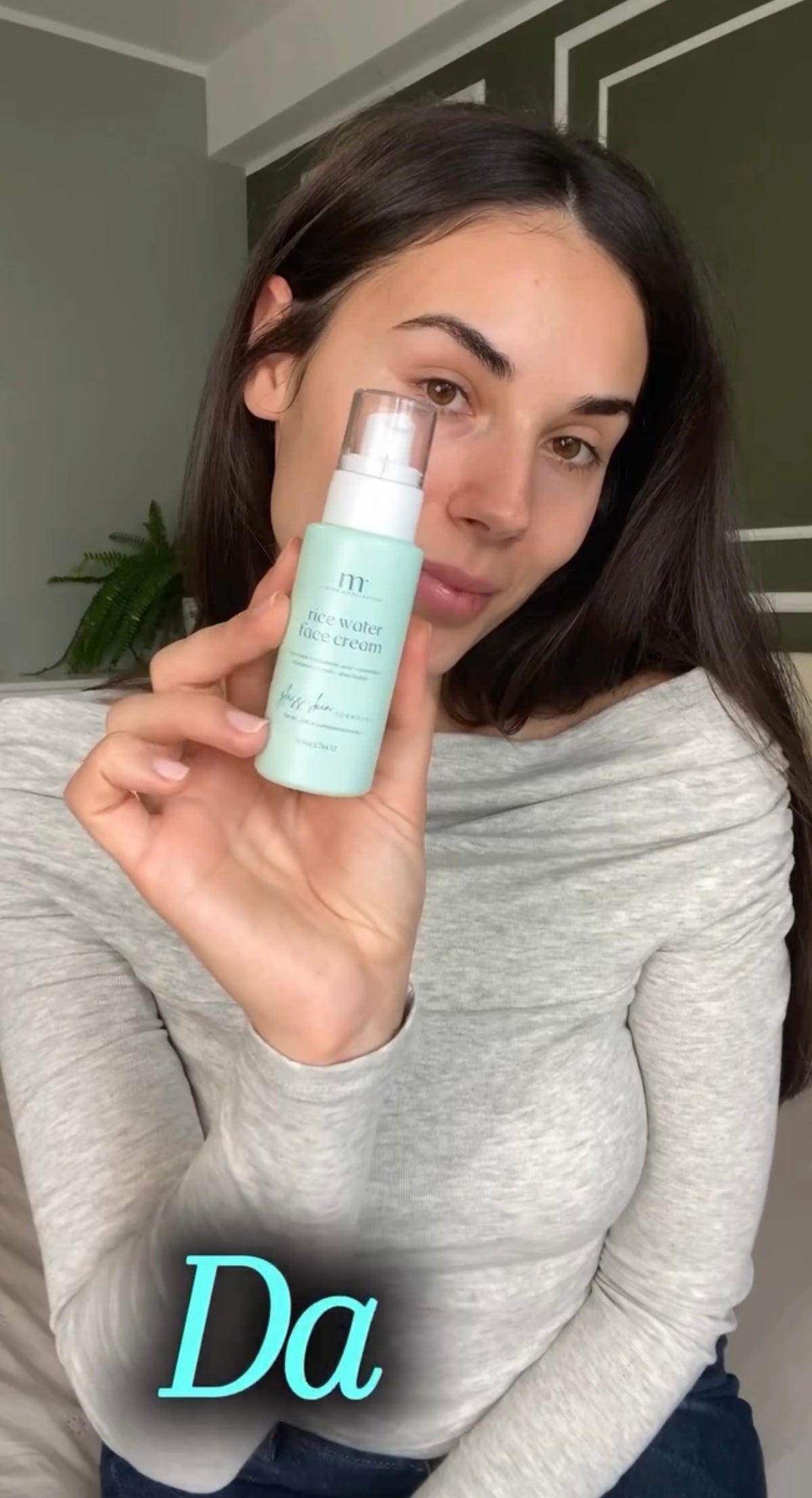 A woman holding up a light mint green cosmetic pump bottle with a clear cap, wearing a gray long-sleeve top. The brand name 'M' is visible on the bottle, and a stylized teal text overlay reading 'Da' is at the bottom.