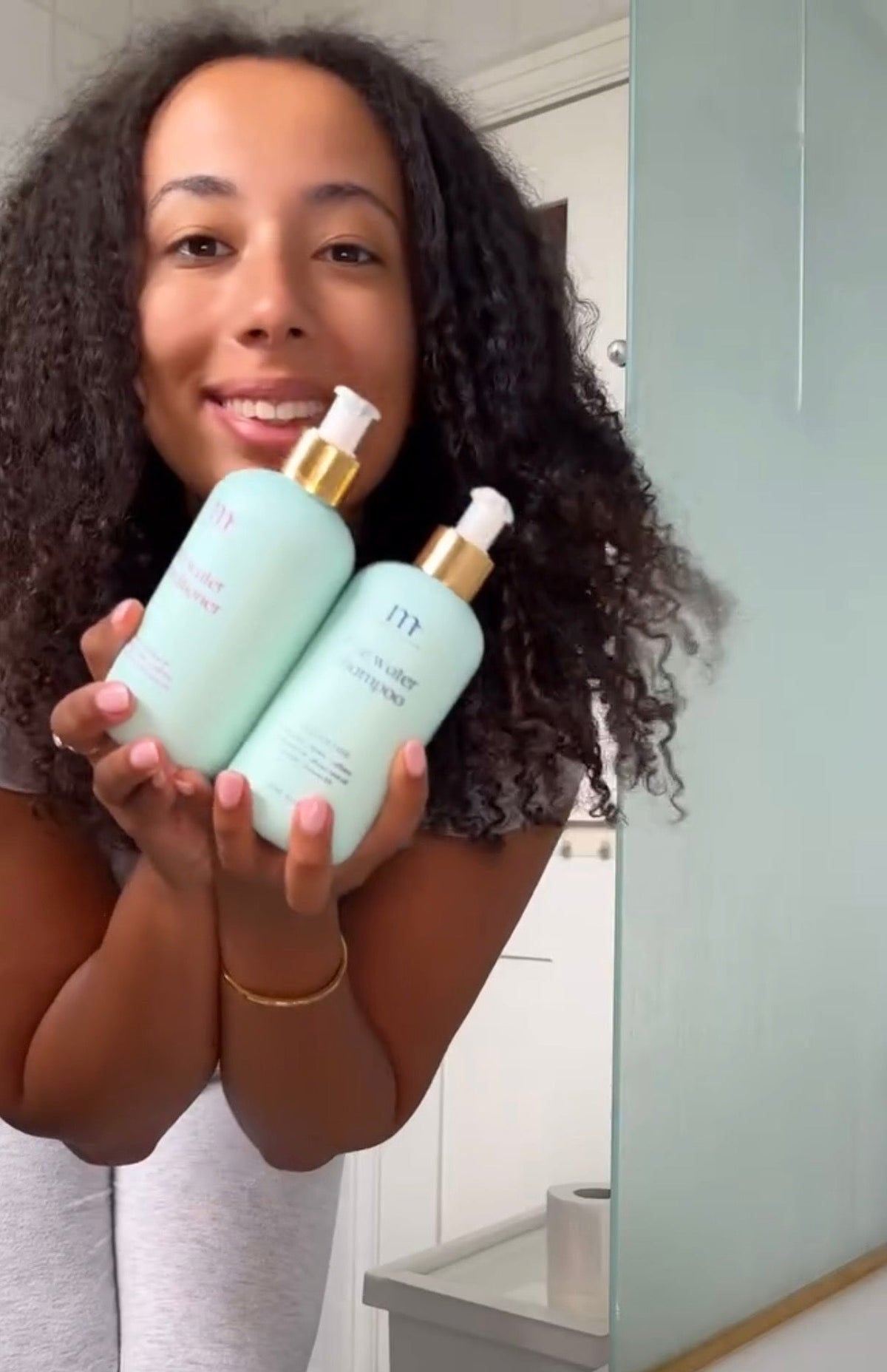 A woman holding two light mint green pump bottles with gold caps in front of a mirror, showing the front of the bottles. The setting appears to be a bathroom with a white tub and a pale green wall.